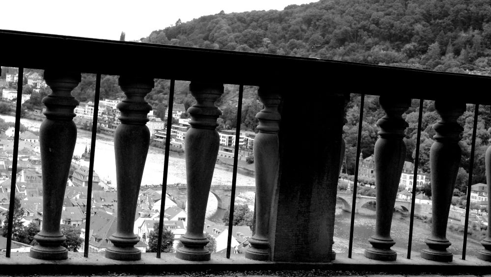 B&w image
The historic balustrade of a castle on a high hill. Through it you can see the Old Bridge, the Neckar and a hill on the other side of the river, trees on it.