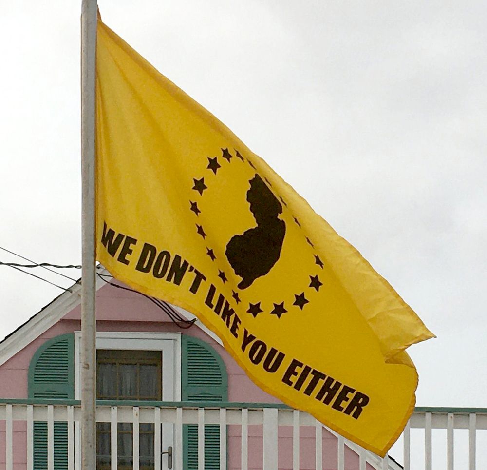 Yellow flag with black images and text. A silhouette of the totally awesome state of NJ ringed by stars, with the text, "We don't like you either" right below it