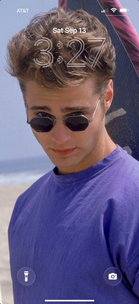 jason priestley, handsome white dude with blue eyes and light brown / dark blond hair, wearing a purple t-shirt looking over a pair of sunglasses at the camera, standing on the beach in front of a purple-and-pink surfboard