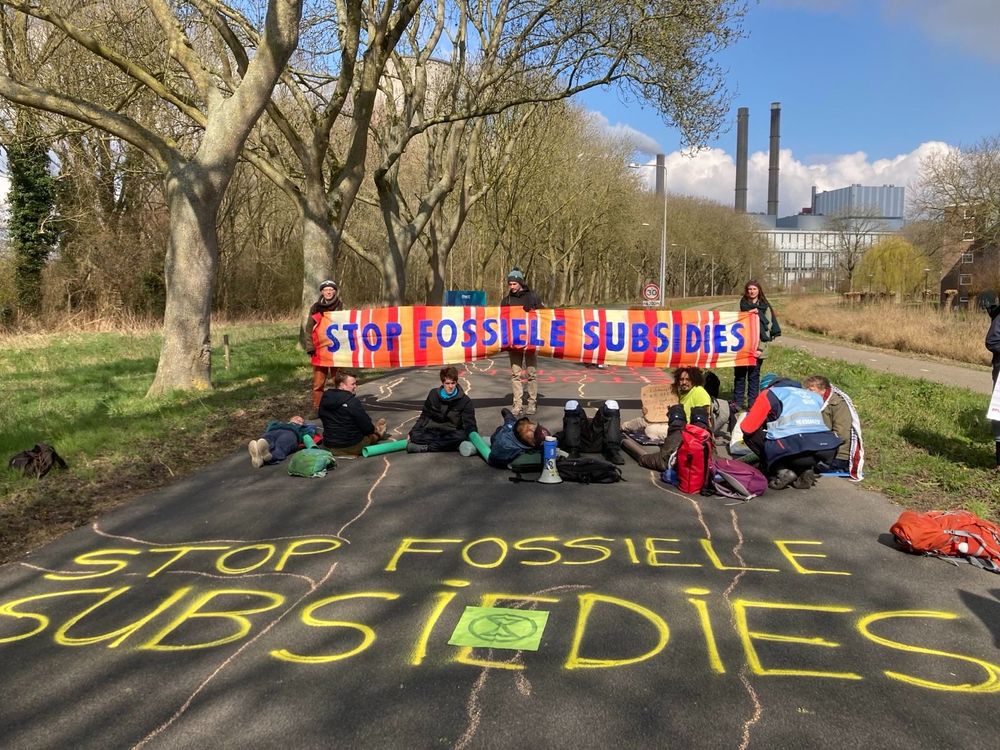 Een groep rebellen blokkeert de weg met spandoeken. De foto is genomen vanaf de voorkant. Op het wegdek voor hun staat met stoepkrijt verschillende leuzen geschreven, zoals stop fossiele subsidies. 