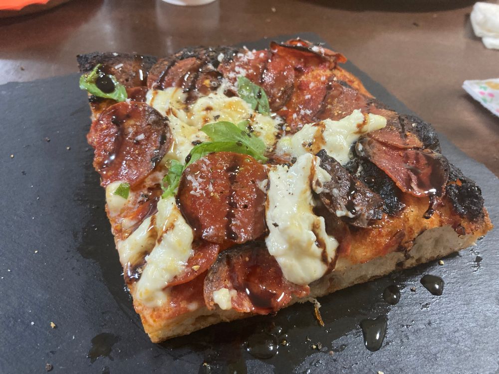 Top down view of a crusty square slice on a charcoal plate, hot honey and balsamic reduction sprayed everywhere