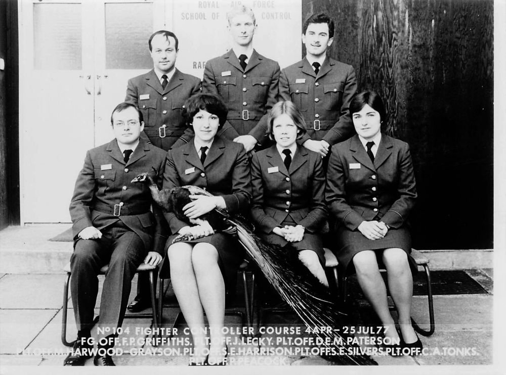 photo, black and white, the No.104 Fighter Controller course. Men and women of the Royal Air Force are seated for a group photo, one lady is holding a peacock on her lap, the peacock is labelled as having the military rank of Pilot Officer.