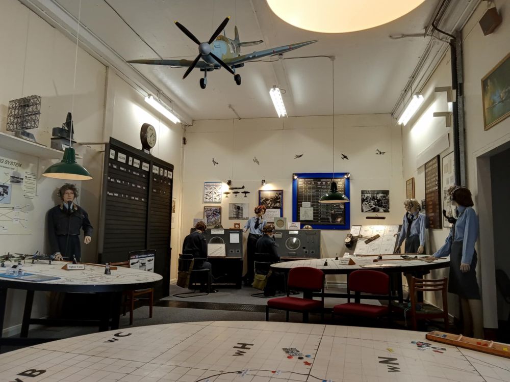 photo, colour, the Second World War room at the RAF Air Defence Radar Museum. The room is painted white, with map tables covered in painted grid squares, mannequins dressed as RAF aircraftswomen, radar consoles, model aircraft and photographs of wartime aircraft.