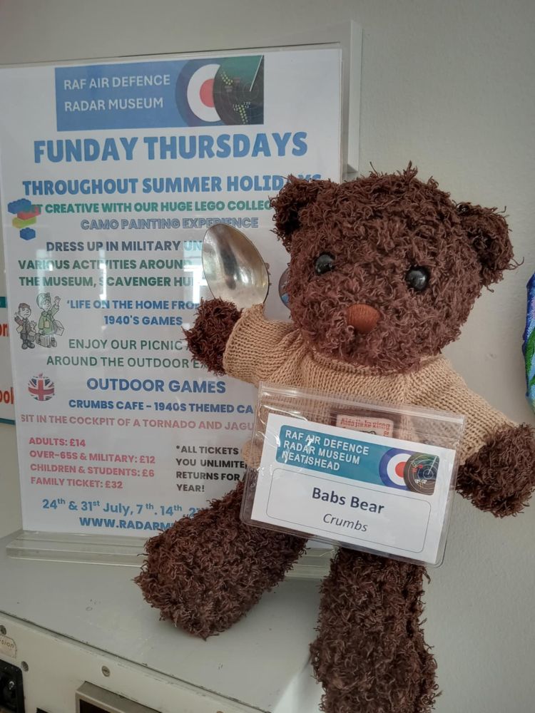 photo, colour, a brown teddy bear wearing a pullover and a name badge stating 'Babs Bear' sitting holding a spoon in front of a poster advertising Funday Thursdays of family events at the RAF Air Defence Radar Museum.