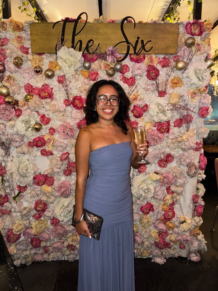 Semi full body picture of me by a flower wall at the six. I have a big smile, a glass of champagne, and my clutch in my hand. 