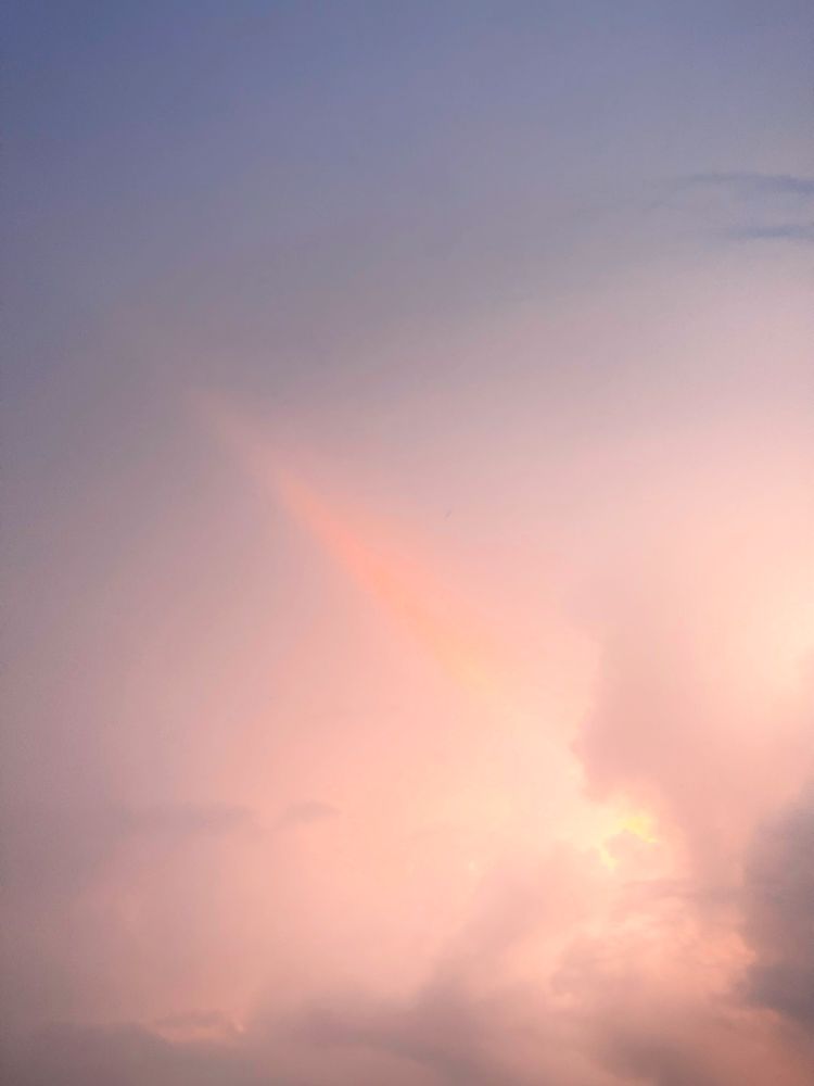 Early dawn sky with clouds shaped and unsettled by Hurricane Erin passing 300 km away. The scene is painted in muted purples, blues, and soft peach light. A delicate diagonal streak of pinkish-orange glow cuts through the cloud layers, like a faint rainbow fragment or beam, while darker cloud masses frame the bottom edge. The overall mood is restless yet serene, capturing the hurricane’s distant turbulence mixed with the calm of morning light.

August 16, Guadeloupe