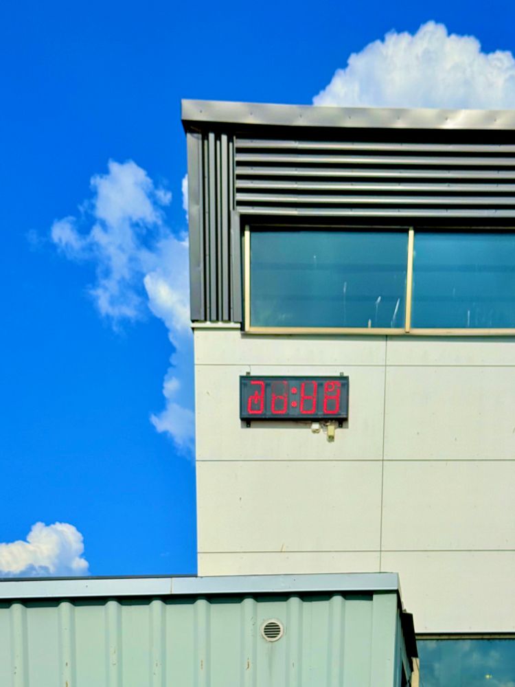 A red LED digital clock is mounted high on the exterior wall of a swimming hall currently undergoing renovation. The clock displays distorted, unreadable digits, suggesting a malfunction or incomplete setup. The building features light gray paneling, vertical ventilation slats, and modern architectural lines, with signs of construction still present. Below, part of a corrugated metal roof—possibly a temporary structure or an entrance canopy—is visible. In the background, a bright blue sky filled with large, white clouds contrasts with the stark, unfinished look of the facility.

July 2025