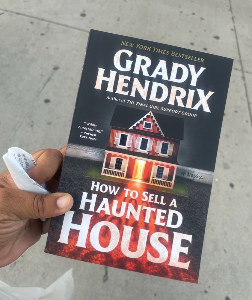 Micro is holding a book with the words How to Sell a Haunted House on the cover. The author’s name “Grady Hendrix” is also in the cover in large font. 