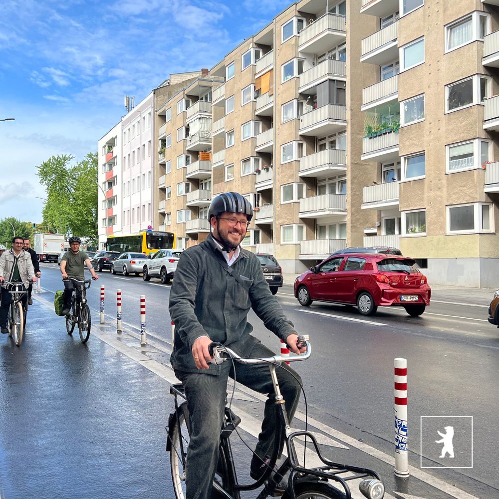 Bezirksstadtrat Christopher Schriner  beim Anradeln auf der Beusselstraße. Der Himmel ist blau. Im Hintergrund sind Wohnhäuser zu sehen. Die Straße ist nass vom Regen.