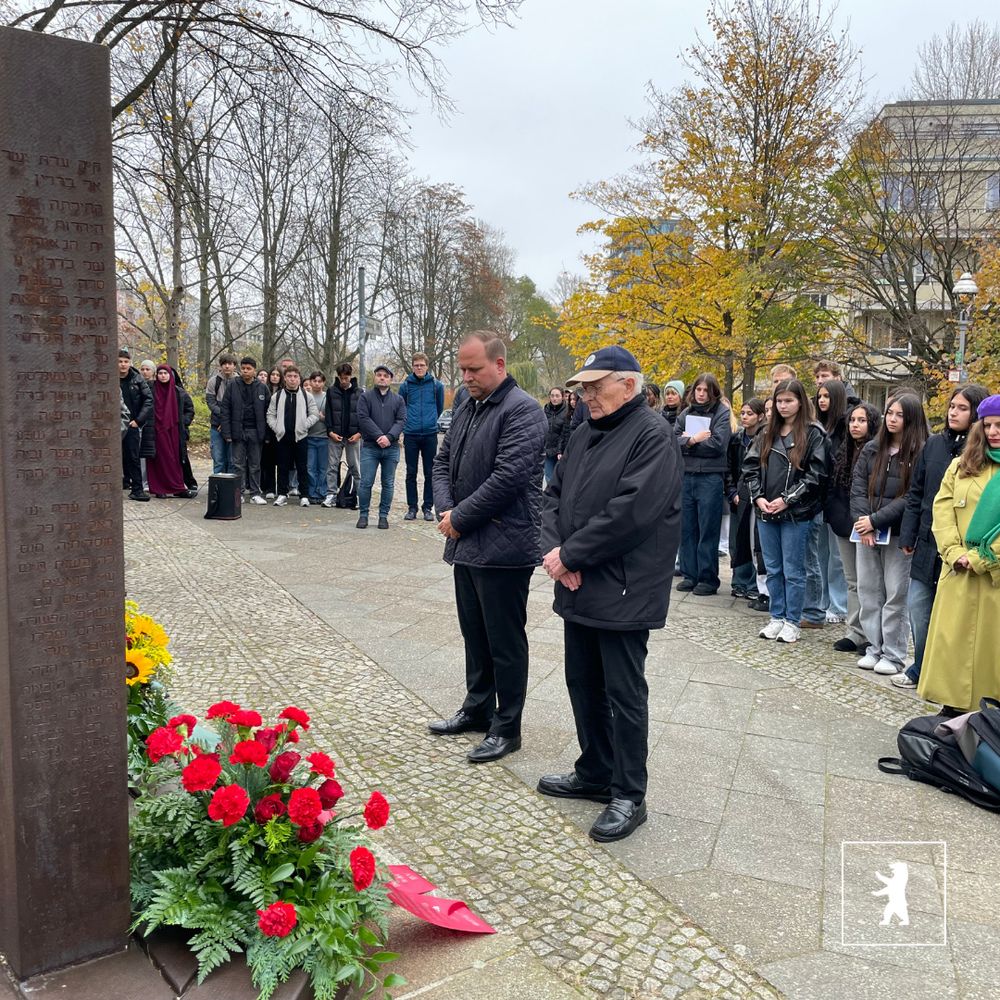 Menschen stehen mit gesenkten Köpfen vor einem Denkmal mit Inschriften, umgeben von Blumenkränzen und Bäumen im Herbst. Unten rechts befindet sich das Logo des BA Mitte.