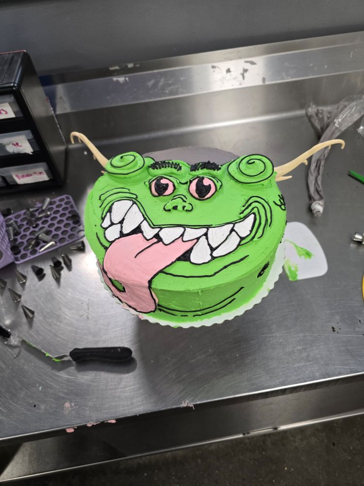 A green horned monster on a round cake. The horns are plastic pieces from a discontinued design. The monster's tongue drops down the side of the cake.