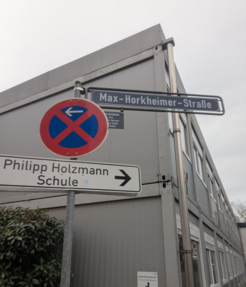 Straßenschild an einer Straßenecke, an der ein grauer Containerbau steht. Es sind drei Schilder angebracht: in der Mitte ein Halteverbotsschild, unten ein Wegweiser zur Philipp Holzmann Schule und ganz oben das Schild mit dem Straßennamen: Max-Horkheimer-Straße (mit zusätzlichem kleinen Hinweisschild, das erläutert, dass Horkheimer ein Vertreter der sog. Frankfurter Schule war).