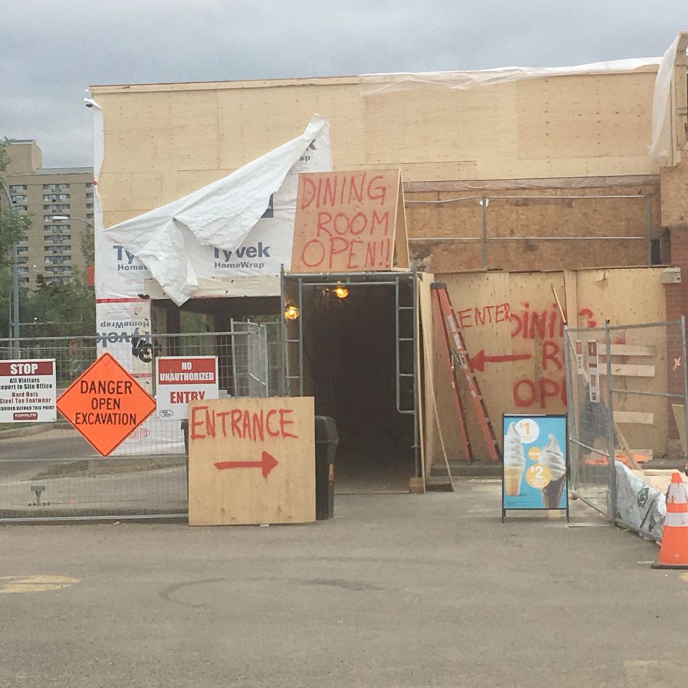 A boarded up McDonald's under construction surrounded by construction signs reading "DANGER OPEN EXCAVATION" and "NO UNAUTHORIZED ENTRY" and spray painted signs in red paint on pieces of plywood saying "DINING ROOM OPEN!!" and "ENTRANCE".