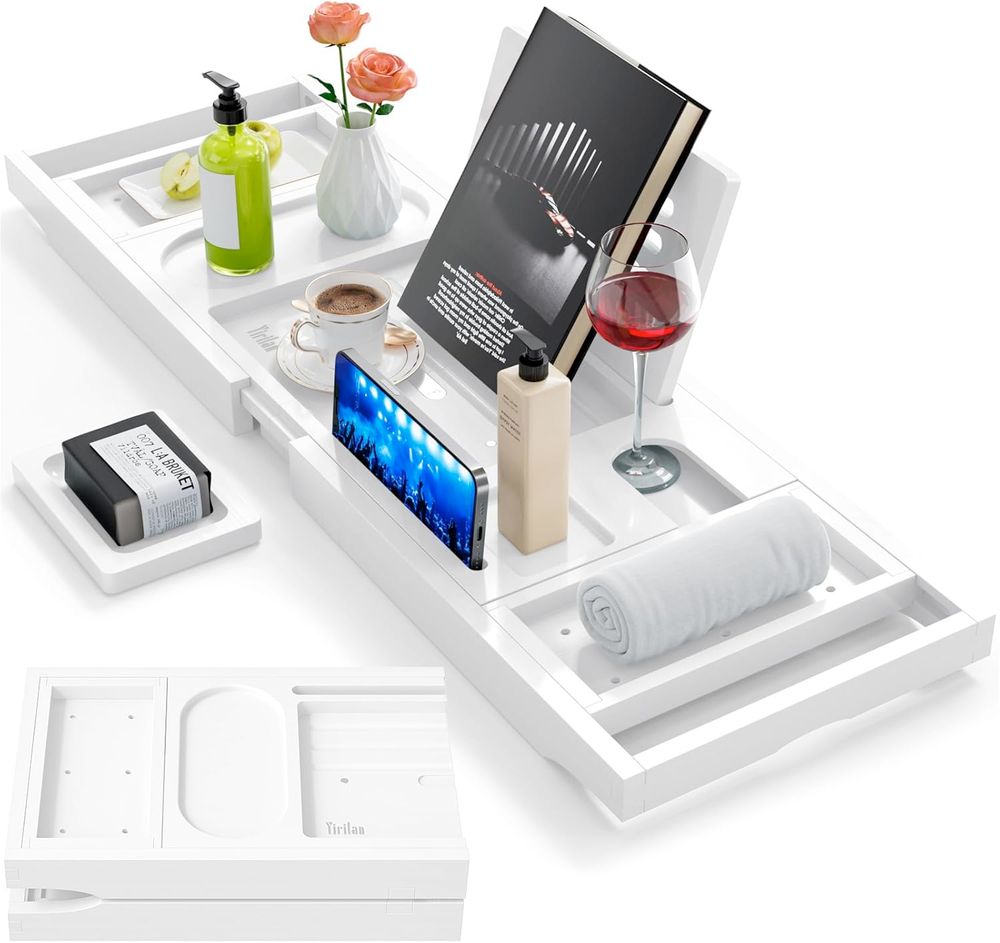 A photo of a white bath try with various items sitting on it including a book, a cellphone, a wine glass, a coffee cup, a thing of lotion, a small towel, a vase with flowers...