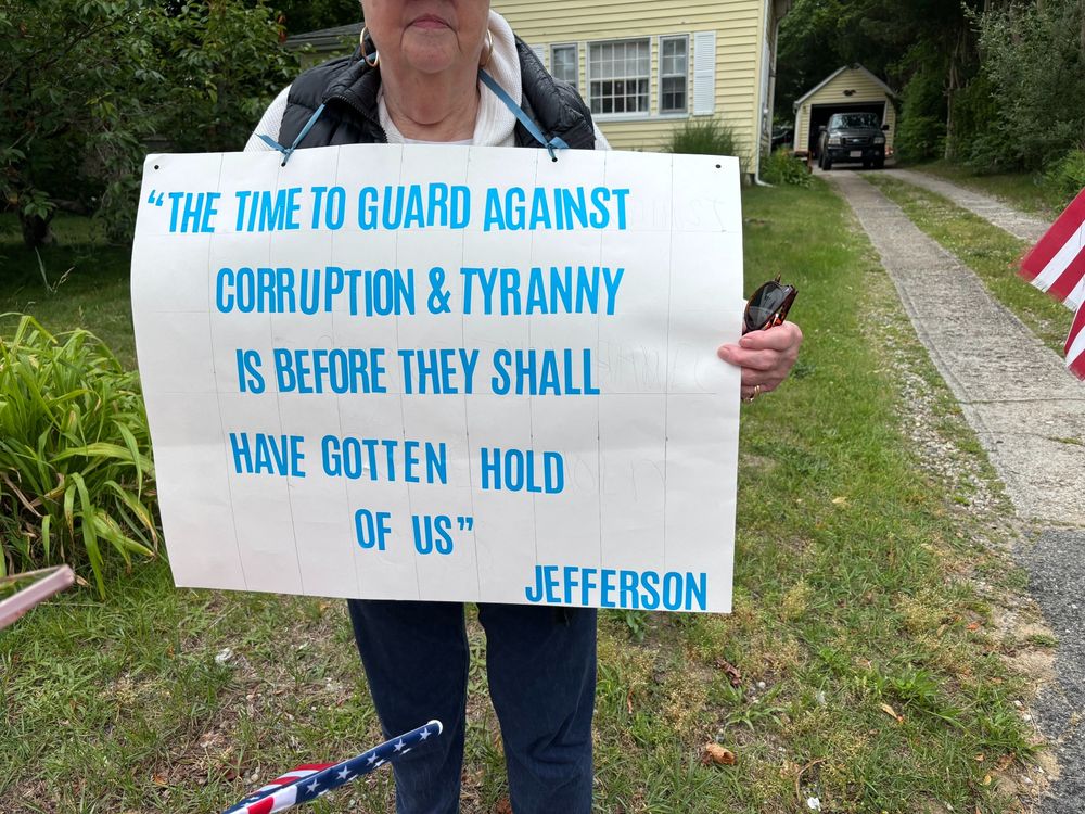 A woman holding a sign with the quotation " The time to guard against corruption & tyranny is before they shall have gotten hold of us" Thomas Jefferson