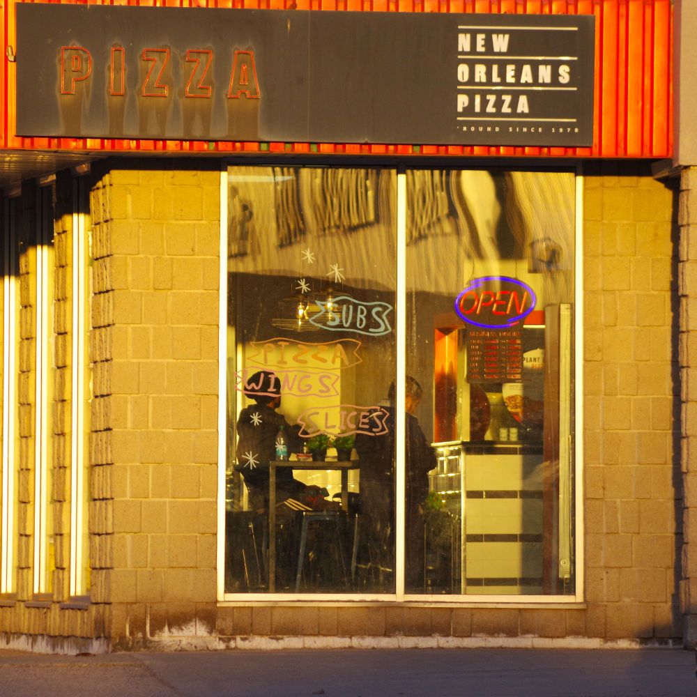 Photograph of a New Orleans Pizza restaurant, bathed in golden hour sunlight.  The glass window is decorated with clumsily drawn words: "Subs" "Wings" "Slices".  There are two people seated inside.