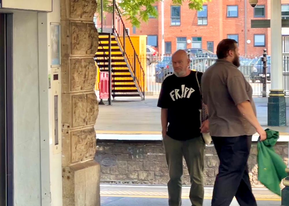 A man on a station platform is wearing a black T shirt with the word Fripp in white lettering