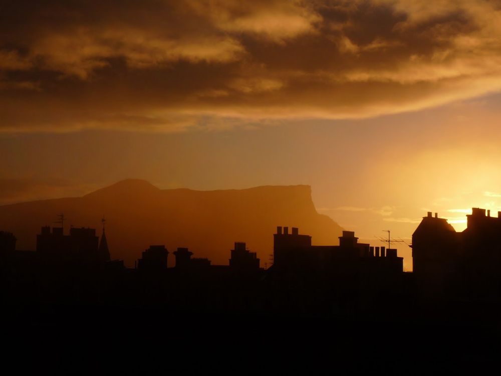A row of rooftops in silhouette as the sun rises. Arthur's Seat glows in the light. A blanket of clouds hover above the promise of a blue sky below 