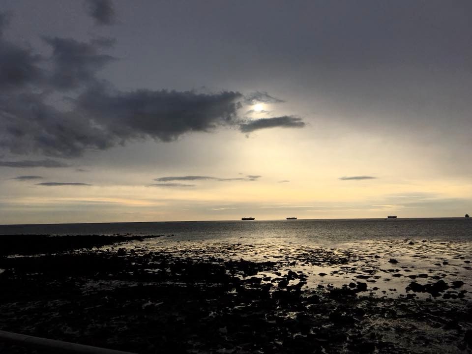 A backdrop of grey cloud cover dominates the sky, while nearer the horizon, a pallid wash of sunlight attempts to brighten the gloom. The shoreline of wet sand reflects the pale light
