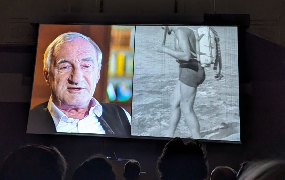 Screenshot from the film with narrator on the left and black and white film on the right 