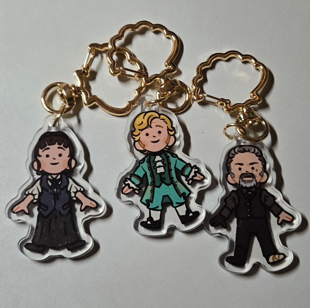 Three keychains of chibi Our Flag Means Death characters: Mary, Stede, and Izzy. They have gold clam shell keyrings.