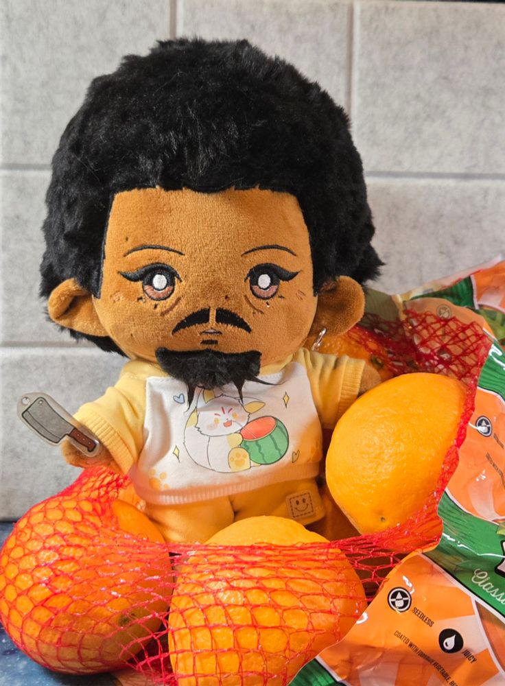 Roach doll standing in a netted bag of naval oranges. He is wearing a shirt with yellow sleeves featuring a cat (maybe a fox?) drinking out of a watermelon through a straw, with a matching pair of yellow shorts. He is brandishing a chef's cleaver made from shrink plastic.