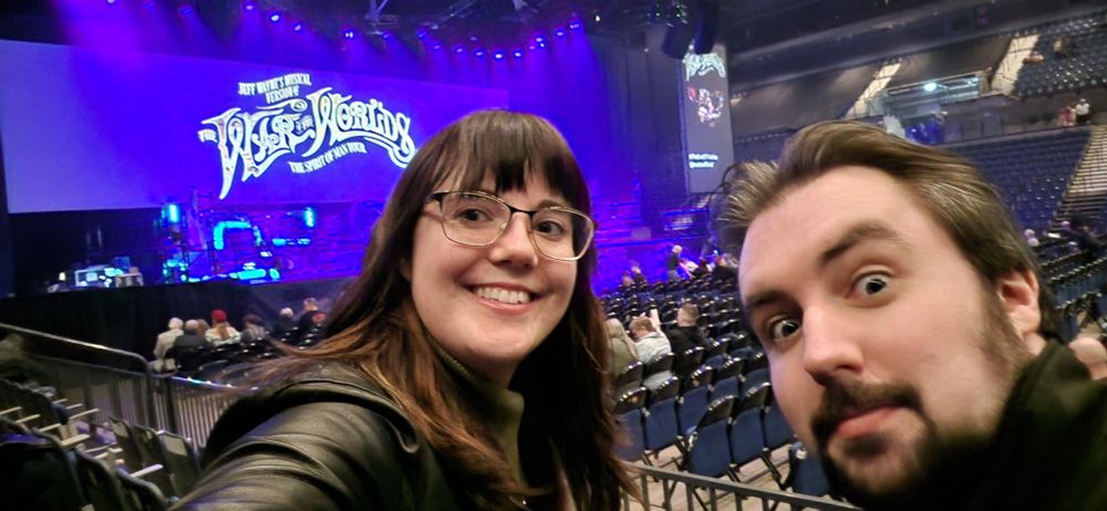 Larissa and Dan smile and make faces to the camera in front of a stage with the words "War of the Worlds" on a screen above