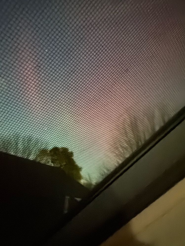 Faint aurora seen through a skylight with a house roof and tops of trees in the foreground. 