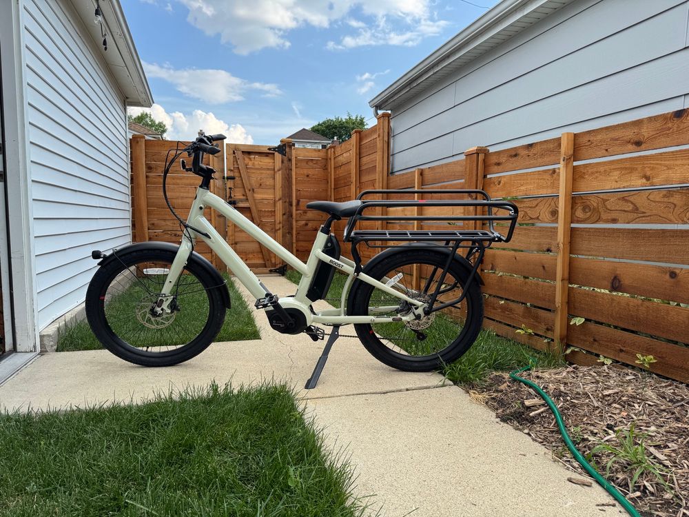 A warm grey colored cargo e-bike with a large rear rack parked on a backyard sidewalk in front of a cedar wood fence. 