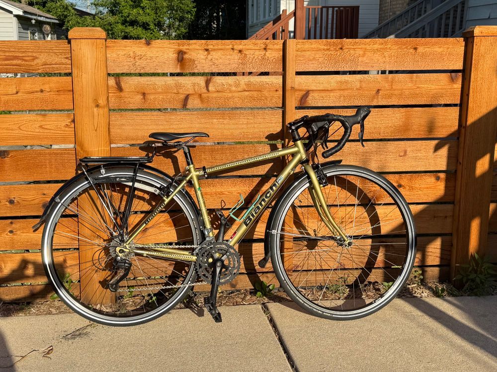 Gold cyclocross bike (Bianchi Volpe) with black pedals, bar tape, saddle, and rack leaning against a cedar wood fence. 