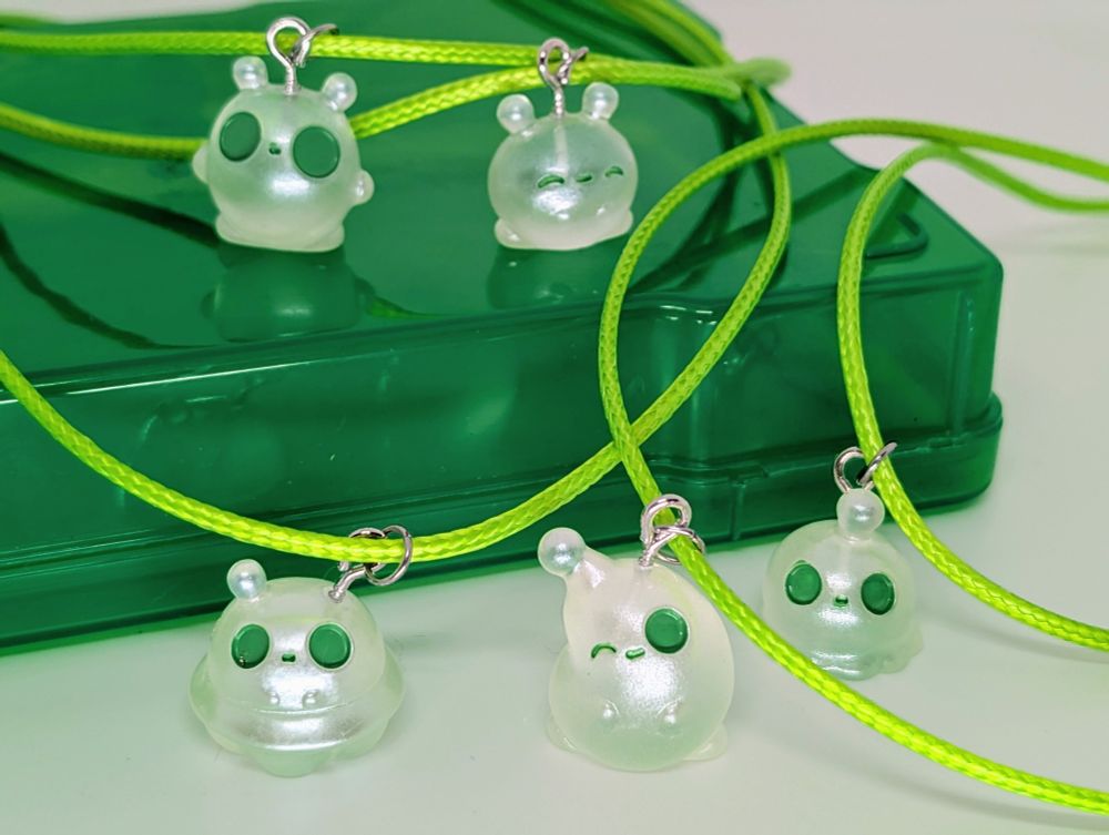 Clear and green alien charms in line green necklaces standing on a green platform with a white background.