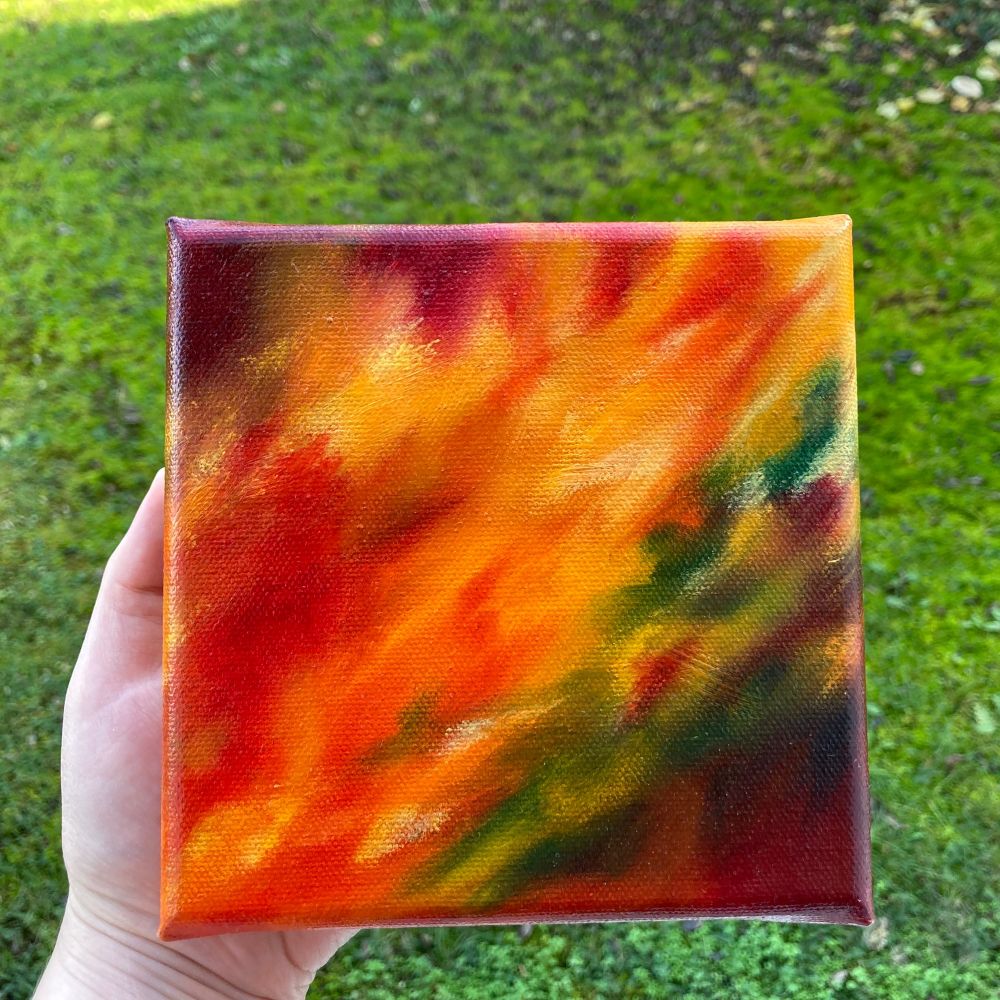 A hand is holding a small abstract oil painting. They colours seem to flow diagonally across the canvas, mostly orange, yellow and bright red in the middle going from the lower left to the upper right. On the lower right there’s some brown and dark red with spots of green, yellow and red on top. There’s another spot of dark red in the upper left corner.