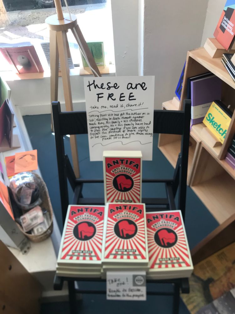 Frame of stack of books with a sign that reads, “these are FREE / take one, read it, share it! / Turning Point USA has put the author on a “list” resulting in death threats against Mark Bray, his family & his students. Consequently, he & his family have had to flee the country. If you are able to support the purchase of more copies so we can continue to give these away, please let us know!”