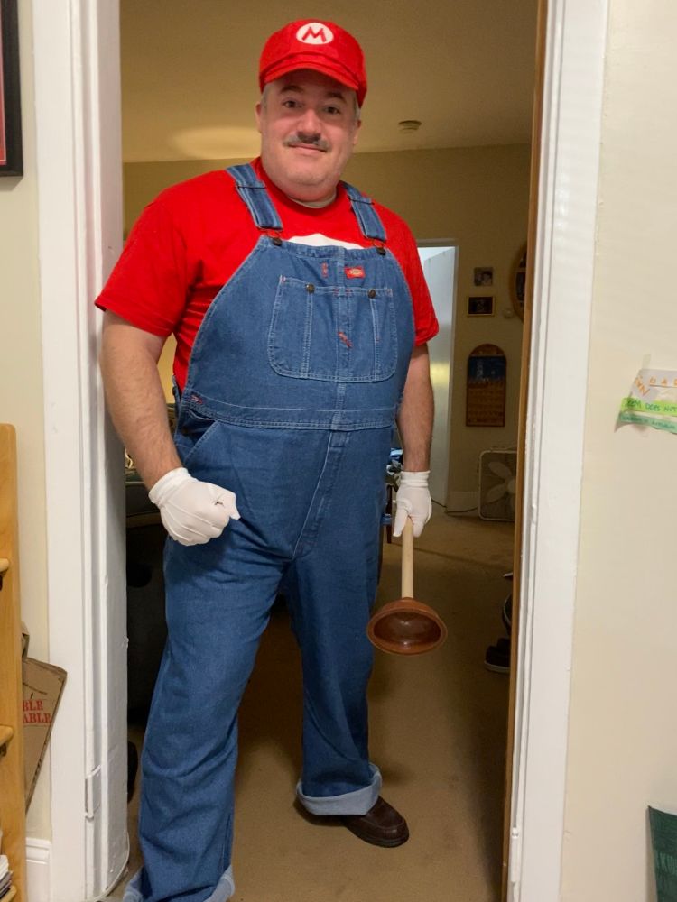 A man with a mustache dressed in blue overalls with a red shirt and red cap bearing an M symbol. He wears white gloves and carries a plunger. 