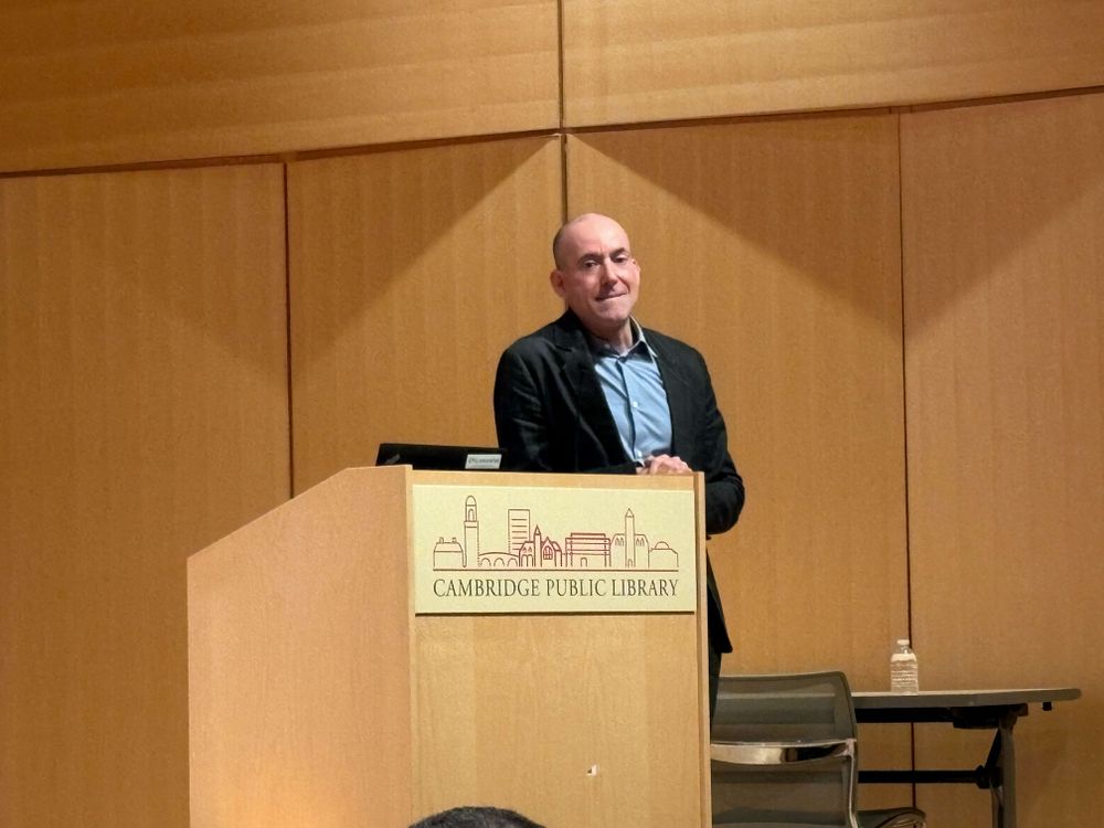 Craig Thompson, a middle-aged Caucasian man, leans agains the Cambridge Public Library podium on stage.