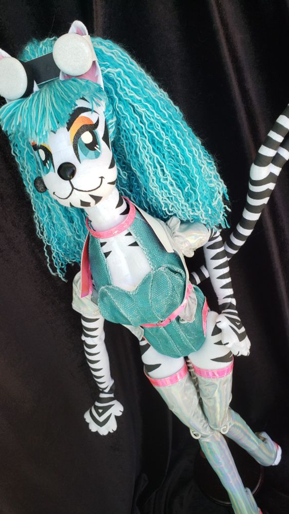 A large cloth doll with blue yarn hair. She is an anthropomorphic feline alien with a long Y-shaped tail. She is made of white cloth and is painted with black stripes. She is painted with bright blue eyes and golden orange eyeshadow. She is wearing a blue bodysuit, a holographic short sleeved jacket and holographic leg warmers. 