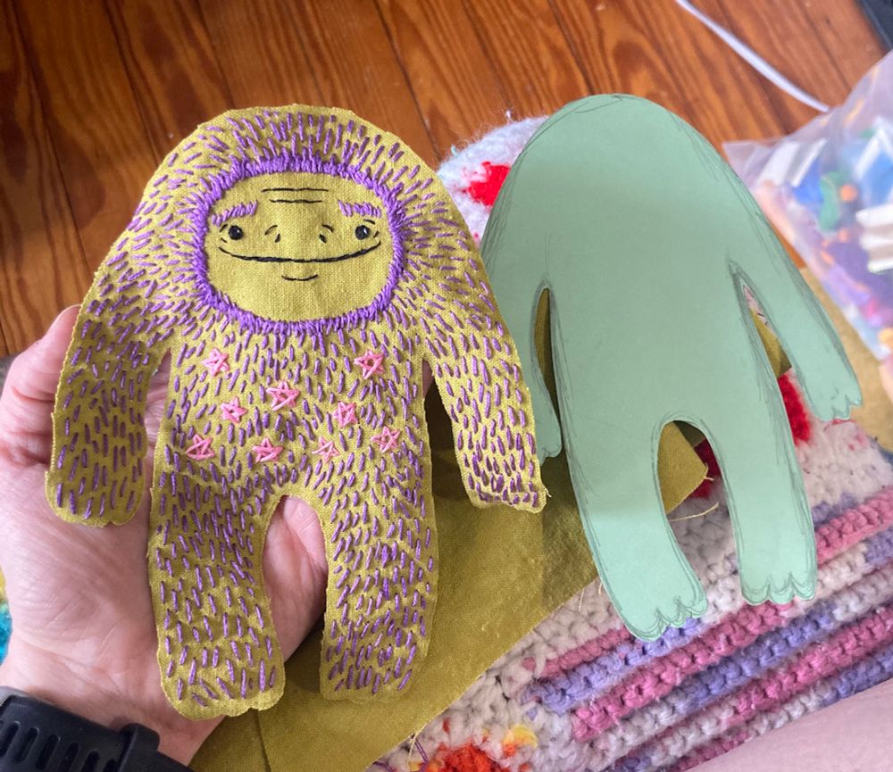 A white person’s hand is holding a hand sewn Sasquatch next to a paper pattern in the same shape. 