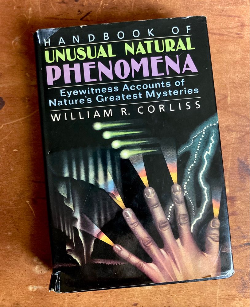 A book sits on a wooden background. The book is titled: “Handbook of Unusual Natural Phenomena: Eyewitness accounts of nature’s greatest mysteries” by William R. Corliss. The cover shows an illustrated hand shooting light fractals from its fingertips. 
