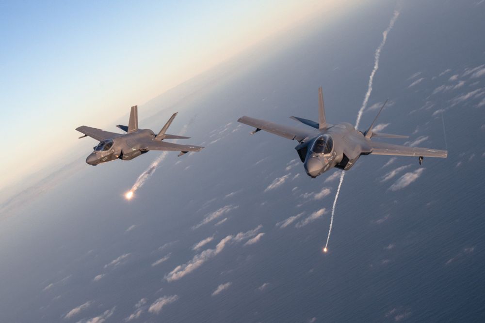Two U.S. Marine Corps F-35C Lightning II stealth fighter jets fly in close formation over the ocean at sunset, with both aircraft releasing bright defensive flares trailing smoke behind them. The jets, marked with U.S. insignia, are part of Marine Fighter Attack Squadrons 311 and 314 participating in Project Legacy, a large-scale training mission conducted off the coast of San Diego on March 4, 2025.