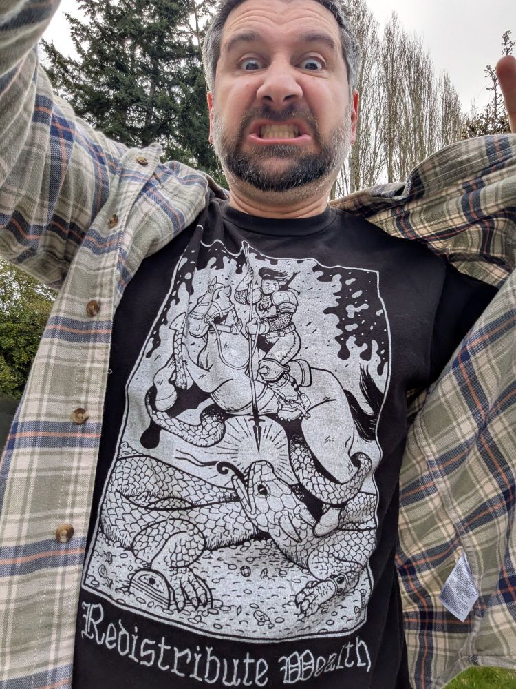 Greg outside with trees behind him grimacing. He's wearing a plaid shirt held open to reveal a black tee shirt with a white print of an Asian-themed warrior slaying a dragon on a bed of gold pieces. The text below reads Redistribute Wealth.
