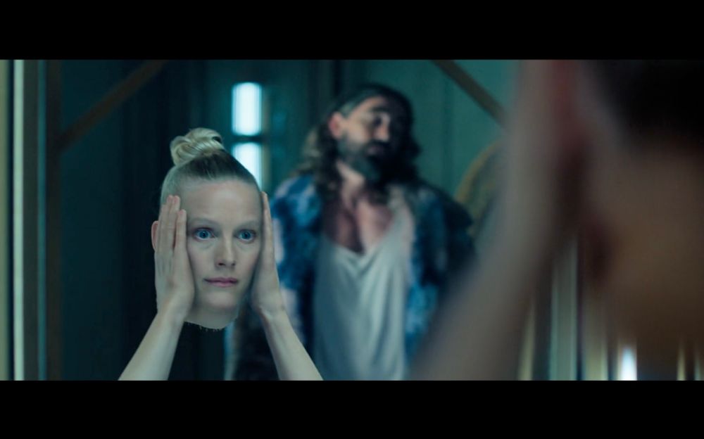 detached woman’s head with it’s hands holding it up while staring into mirror with man in background 