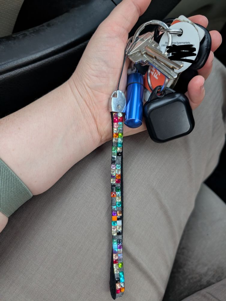 A left hand holding a keychain set over a pair of slacks. There are too many keys on it. And various chunky keychains. It probably weighs more than a tiny could carry. There's also a long dangling rhinestone-covered lanyard.
