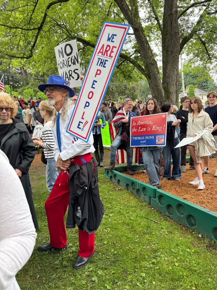Cool older man dressed as an All American Uncle Sam with pimp styling including red slacks and a blue fedora, holding a red white and blue sign that reads “WE ARE THE PEOPLE”
