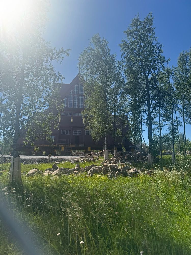 A large wooden church is sitting on yellow metal girders. In the foreground is a grassy area. The photo is taken through a chain link fence, which can be seen in the picture 