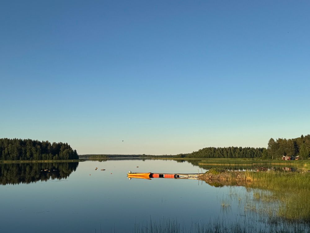 A cloudless sky over still lake water, with a few rowboats in it, rounded by pine forest 
