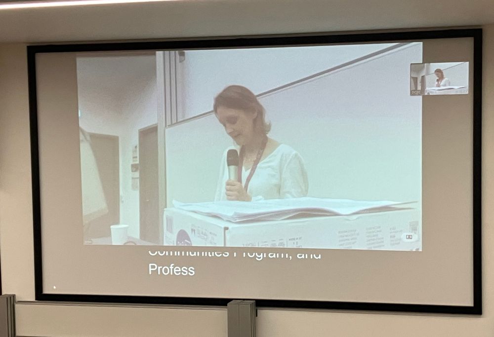 Photo of the large screen in lecture theatre with captions. The first line is cut in half by the video feed