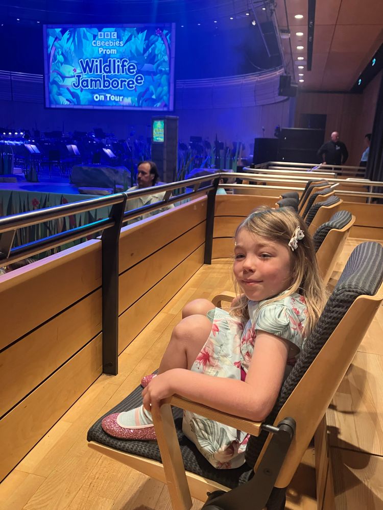 Small blonde child sits in theatre style seats with screen in background reading Wildlife Jamboree 