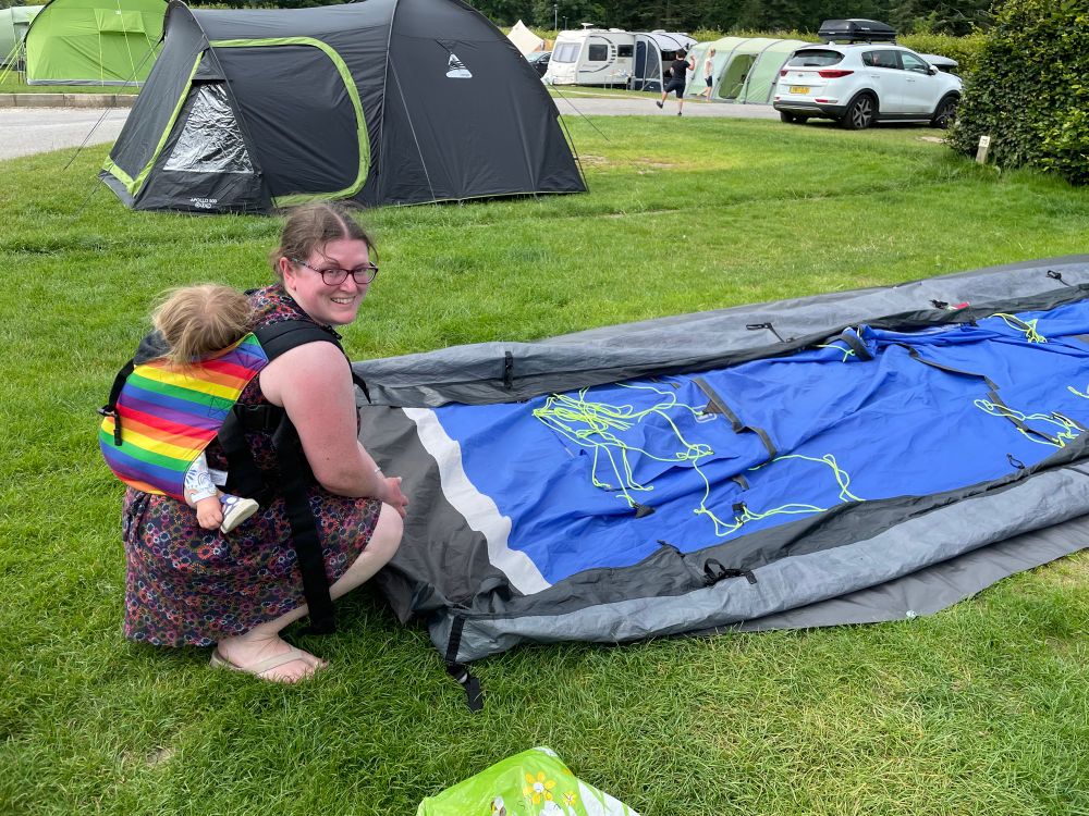 Woman crouching on grass with flattened tent with toddler in rainbow connecta sling on back asleep 