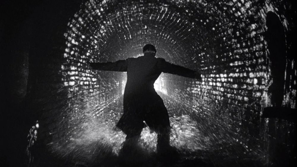 B&W shot of a man standing in a sewer,  silhouetted by light at the end of the tunnel