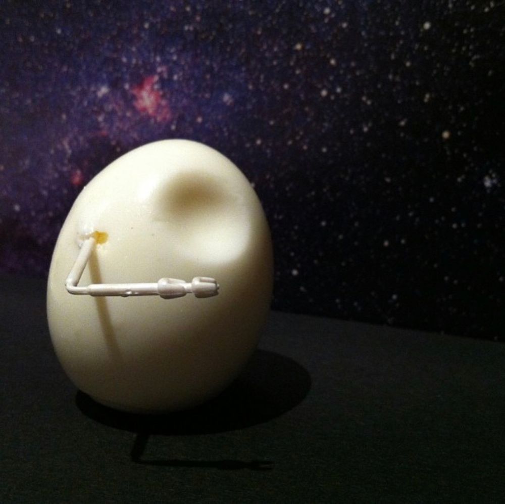 A boiled egg with a circular dent hear the top. It is  photographed in front of an open book which has a picture of a star field. There is a piece of thin bent plastic attached to it, resembling an arm. The overall effect is to make the egg look like an EVA pod from 2001: A Space Odyssey 